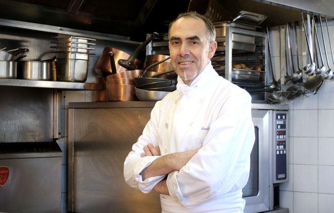 Chef Bruno Seillery souriant bras croisés dans une cuisine professionnelle avec des casseroles, poêles et ustensiles suspendus en arrière-plan