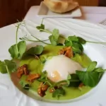 Assiette blanche avec une préparation gastronomique composée d'un œuf poché au centre, entouré d'une sauce verte, de pousses de plantes vertes et de morceaux de champignons dorés, avec un morceau de pain en arrière-plan sur une table blanche.