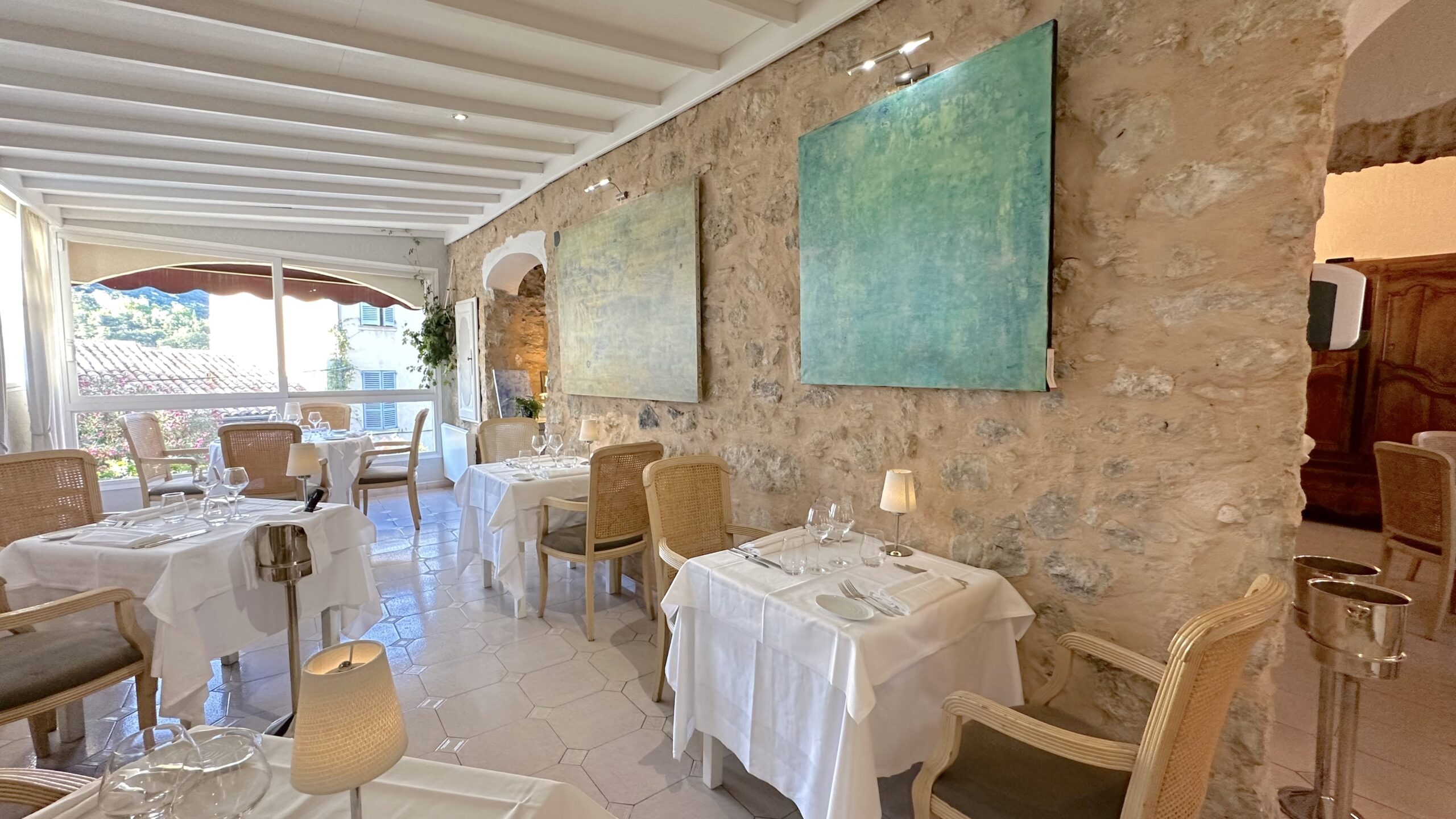 Intérieur du restaurant élégant avec des murs en pierre apparente et des tables dressées avec nappes blanches, verres et couverts, éclairage doux avec des lampes sur les tables et de grandes fenêtres laissant entrer la lumière naturelle.