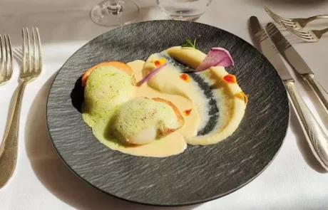 Plat gastronomique présenté sur une assiette noire avec deux portions de coquilles Saint-Jacques nappées de sauce verte et crème, accompagnées de purée décorée de garnitures colorées, le tout posé sur une table avec couteaux, fourchettes et verres à vin.