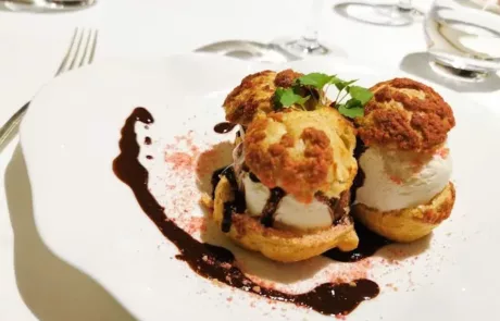 Deux choux à la crème garnis de glace et nappés de sauce au chocolat, décorés de feuilles vertes sur une assiette blanche avec une table dressée en arrière-plan.