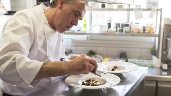 Bruno Seillery décorant minutieusement des plats sur deux assiettes dans une cuisine professionnelle lumineuse.