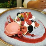 Dessert sur une assiette grise avec une quenelle de sorbet rose, des figues coupées, des fruits rouges variés, de la chantilly et un coulis rouge décoratif.