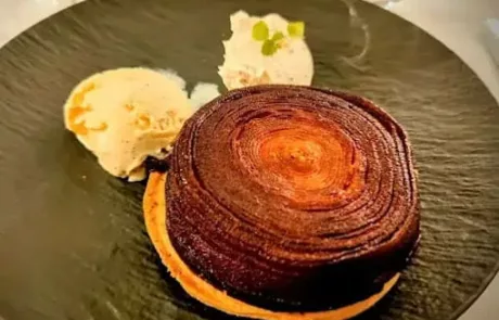 Tartelette au chocolat avec une base biscuitée, accompagnée d'une boule de glace à la vanille et d'une petite garniture blanche décorée de feuilles vertes, le tout présenté sur une assiette noire texturée.