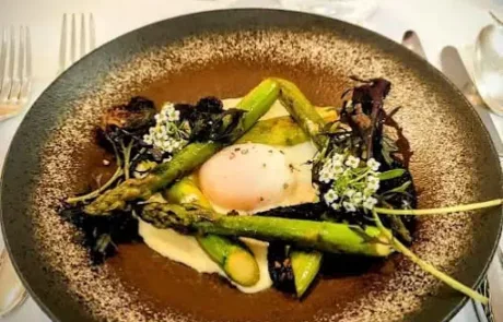 Assiette gastronomique avec asperges vertes, œuf poché, purée blanche et garniture de feuilles et fleurs comestibles sur une table avec couverts et verre en arrière-plan.
