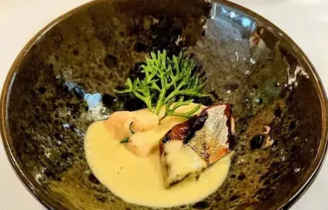 Plat gastronomique dans un bol noir, composé d'un morceau de poisson grillé nappé d'une sauce crémeuse jaune, accompagné de quelques légumes verts et blancs, sur une table avec un verre d'eau en arrière-plan