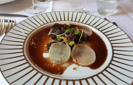 Assiette blanche à motifs rayés avec un plat gastronomique de morceaux de viande en sauce foncée, garnis de fines tranches de radis et de pousses vertes, sur une table avec nappe blanche et couverts en argent.