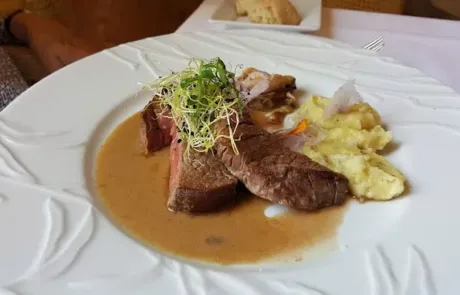 Plat gastronomique avec tranche de viande accompagnée de purée, garnie de pousses vertes et nappée d'une sauce crémeuse sur une assiette blanche au design en relief.