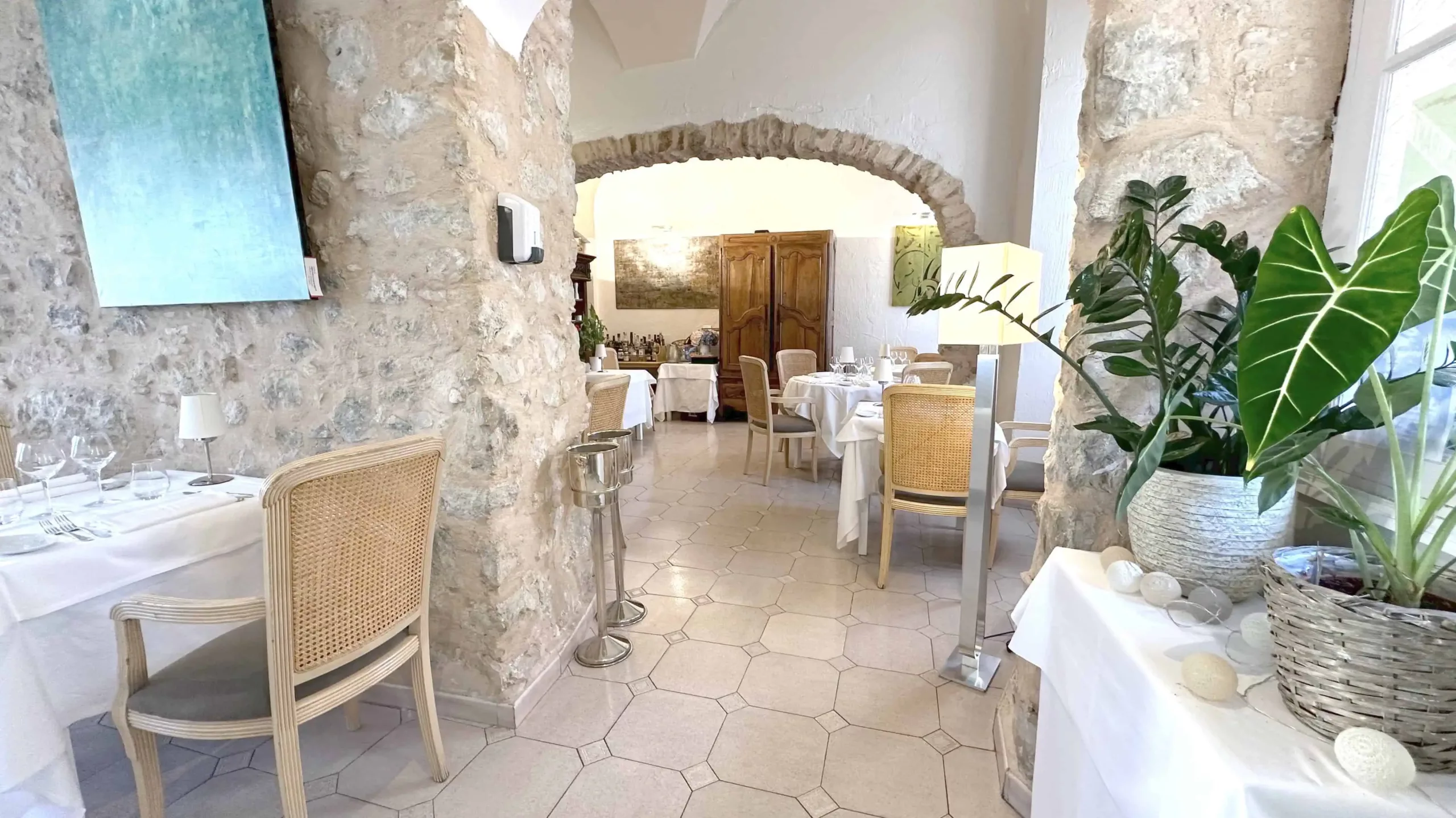 Salle de restaurant avec murs en pierre, tables dressées avec nappes blanches, chaises en bois clair et osier, décorations végétales en pots, et un meuble en bois au fond sous une arche en pierre.