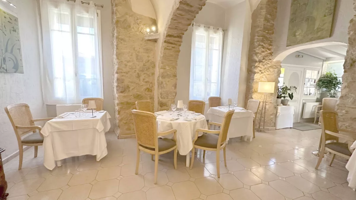 l’Ambroisy – restaurant gastronomique Vence (1) Salle de restaurant lumineuse avec murs en pierre et arches, tables rondes et carrées nappées de blanc, chaises en bois clair avec assise grise, grandes fenêtres avec rideaux blancs, lampadaire et plantes vertes en arrière-plan.